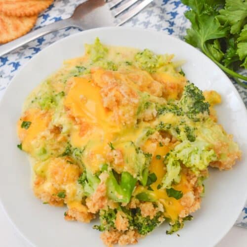 A plate of Velveeta Broccoli Casserole, topped with melted cheese and golden breadcrumbs, sits next to fresh parsley, crackers, and a fork on a patterned cloth.