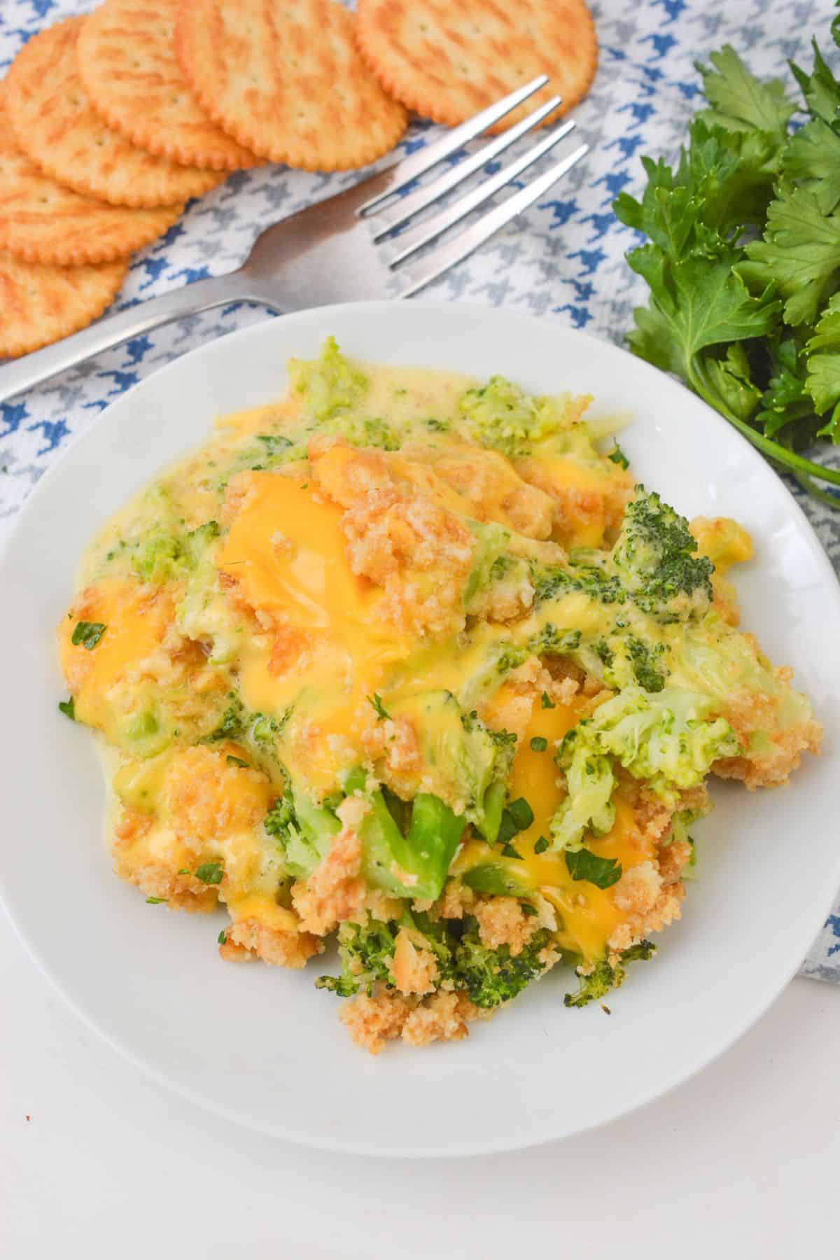 A plate of Velveeta Broccoli Casserole, topped with melted cheese and golden breadcrumbs, sits next to fresh parsley, crackers, and a fork on a patterned cloth.