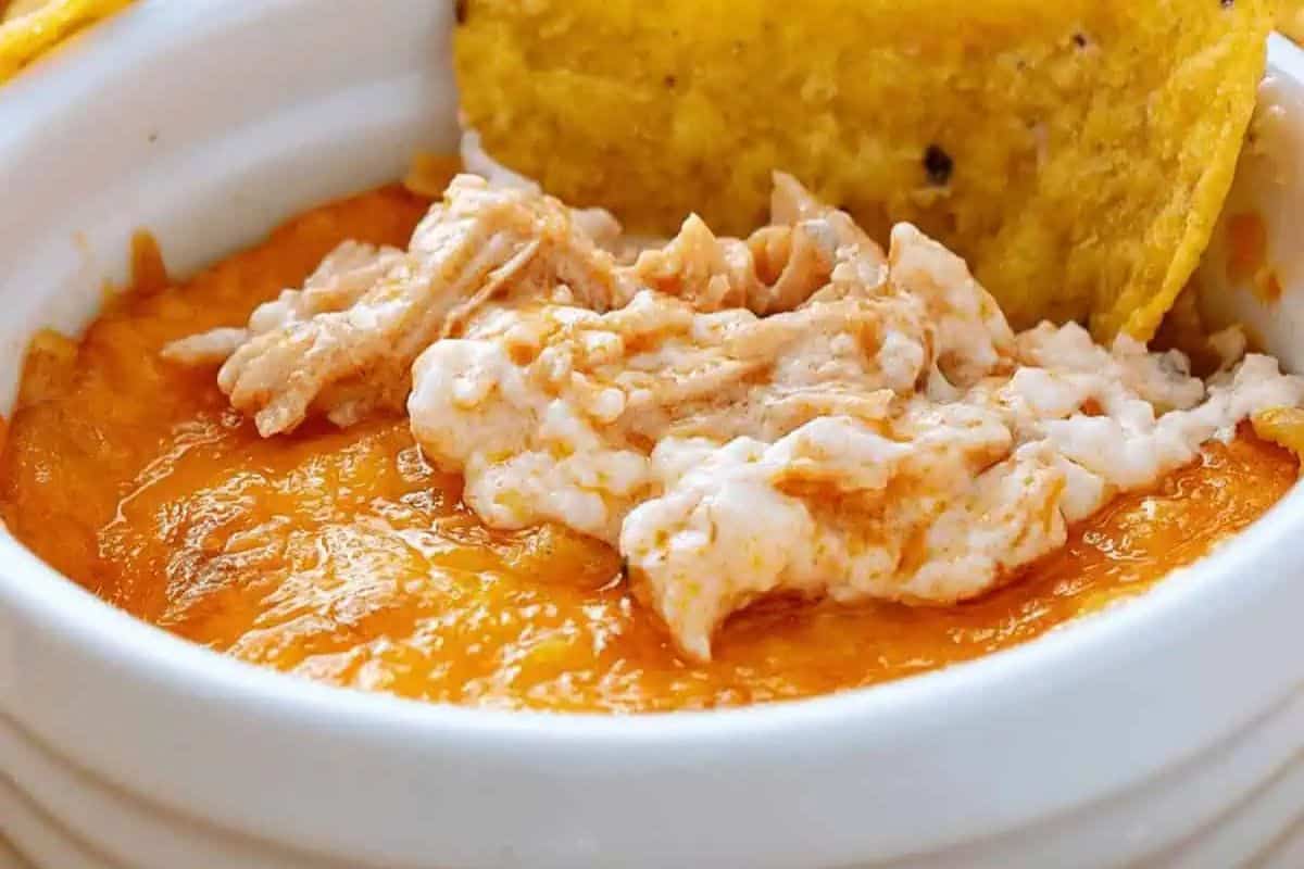 Close-up of a baked cheesy dip topped with shredded chicken in a white dish, with a yellow tortilla chip dipped in—perfect for serving as patriotic party food at your next Labor Day or Memorial Day celebration.