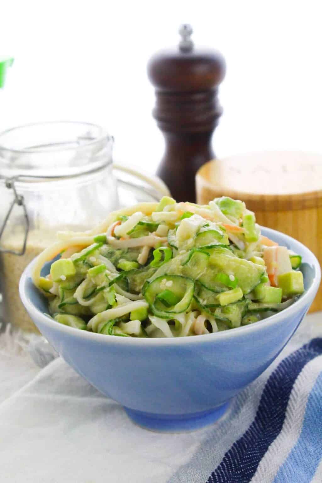 A blue bowl filled with creamy pasta and vegetables, inspired by California Roll Cucumber Salad, sits on a cloth next to a jar, a pepper grinder, and a wooden salt container.