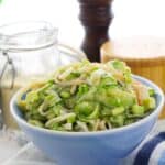 A blue bowl filled with creamy pasta and vegetables, inspired by California Roll Cucumber Salad, sits on a cloth next to a jar, a pepper grinder, and a wooden salt container.