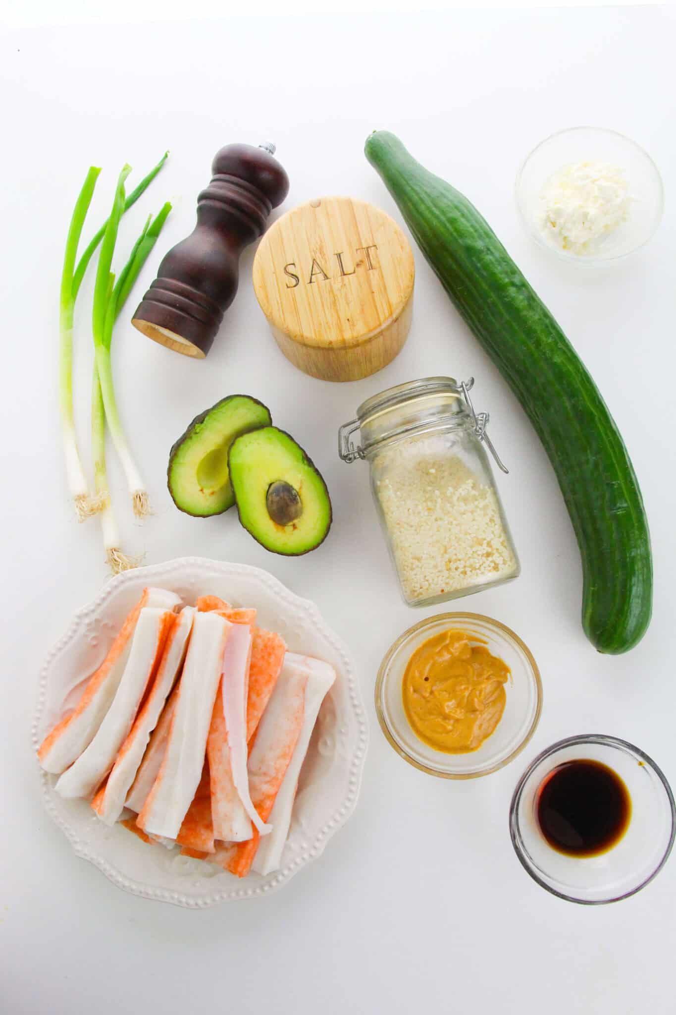 Ingredients for a California Roll Cucumber Salad on a white background, including imitation crab sticks, avocado, cucumber, green onions, rice, soy sauce, wasabi, mayonnaise, salt, and pepper.