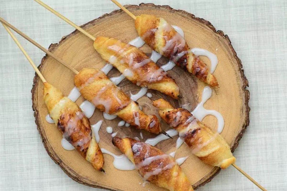 Flavor-packed, pastry-wrapped treats on wooden skewers are arranged on a round wooden board, drizzled with white icing.