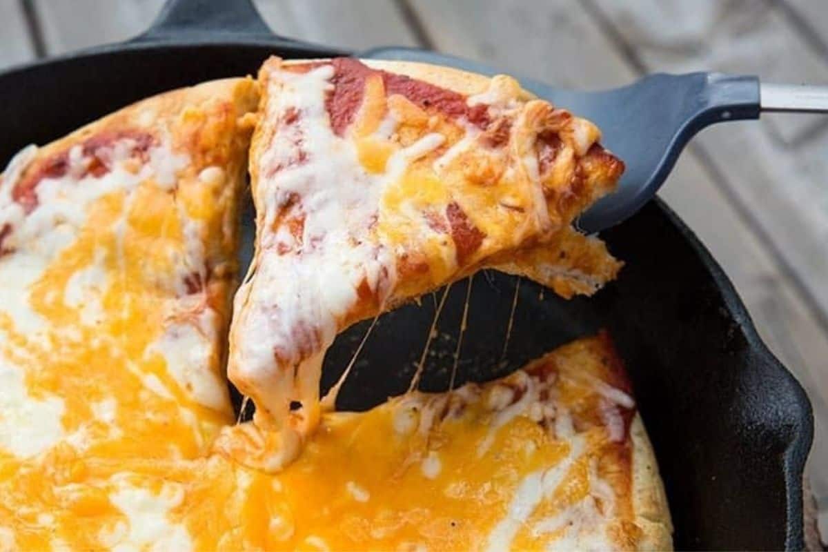A spatula lifts a flavor-packed, cheesy slice of pizza with a thick crust from a pan, while melted cheese stretches from the remaining pizza.
