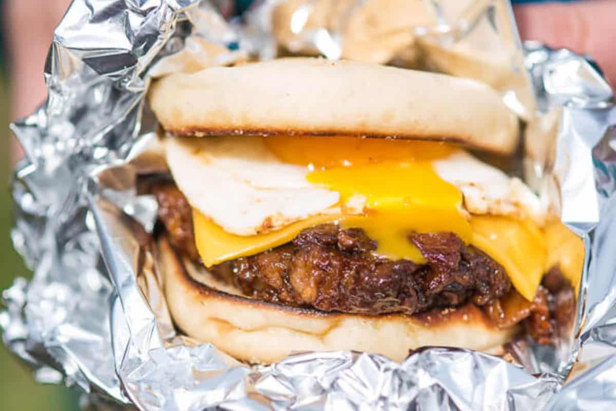 A close-up of a flavor-packed breakfast sandwich with a fried egg and melted cheese, served on a bun and wrapped in aluminum foil.