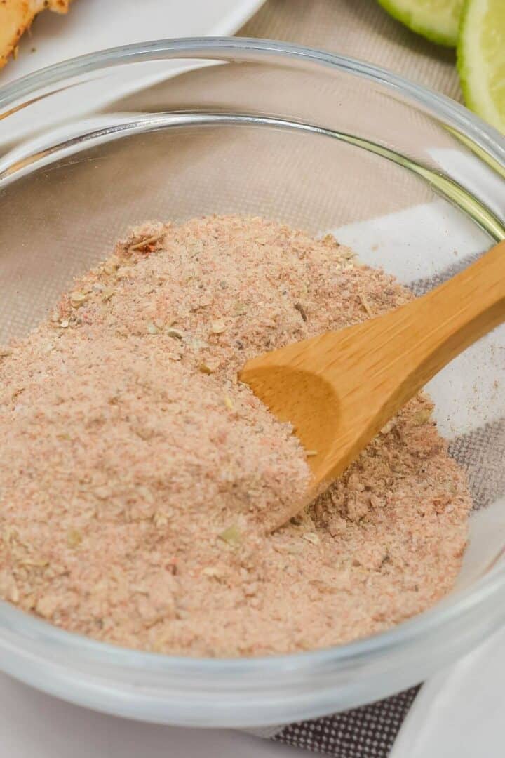 A glass bowl filled with a light brown, finely ground carnitas seasoning, with a wooden spoon partially submerged in the mixture.
