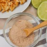 A glass bowl of mix with a wooden spoon sits next to sliced cooked chicken breast and halved limes on a table.