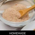 A glass bowl filled with flavorful carnitas seasoning, with a wooden spoon resting inside. Plates of food are visible in the background.
