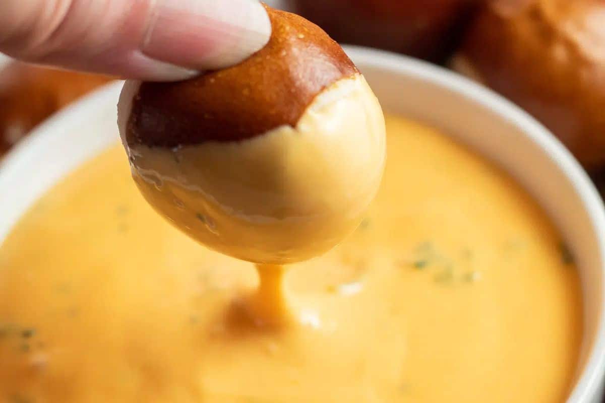 A hand dips a pretzel bite into a bowl of creamy cheese sauce, bringing German favorites to life—perfect for your next Oktoberfest or as one of your easy recipes.