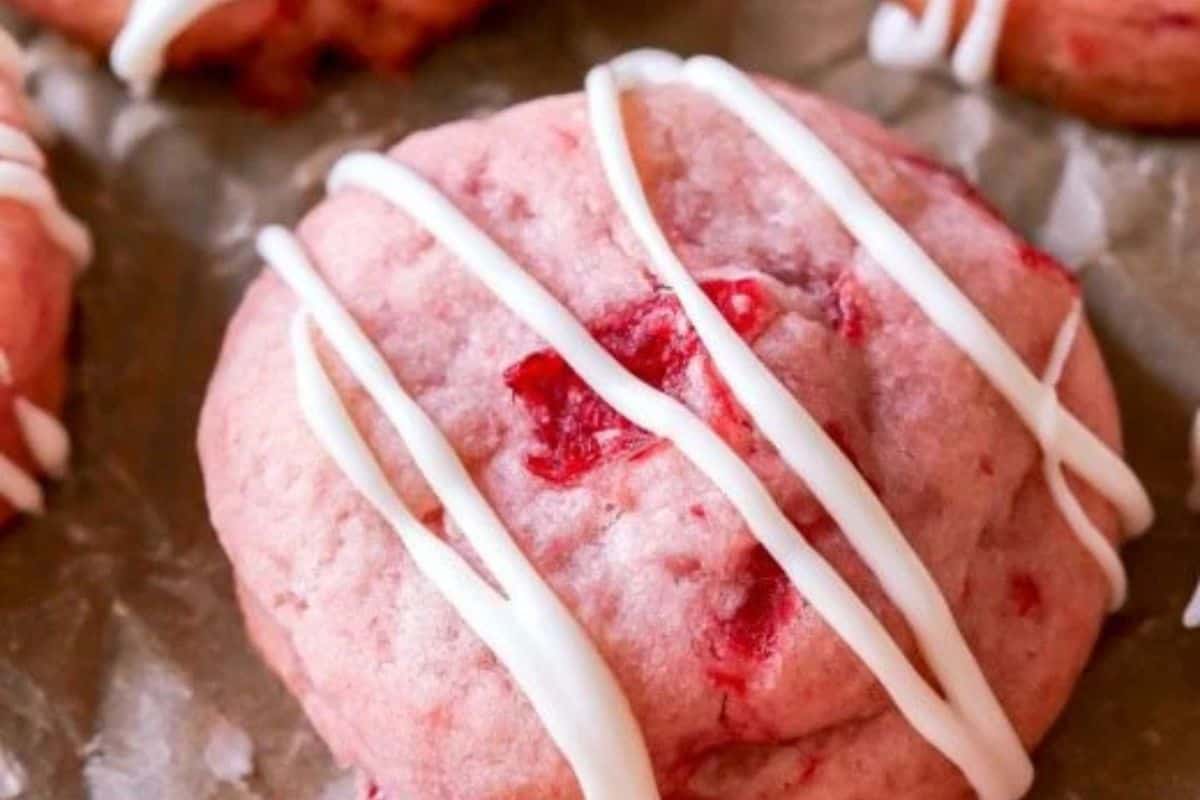 A close-up of a pink cookie with visible red fruit pieces, topped with white icing drizzle, resting on parchment paper—a delightful treat perfect for those who love cherry desserts.