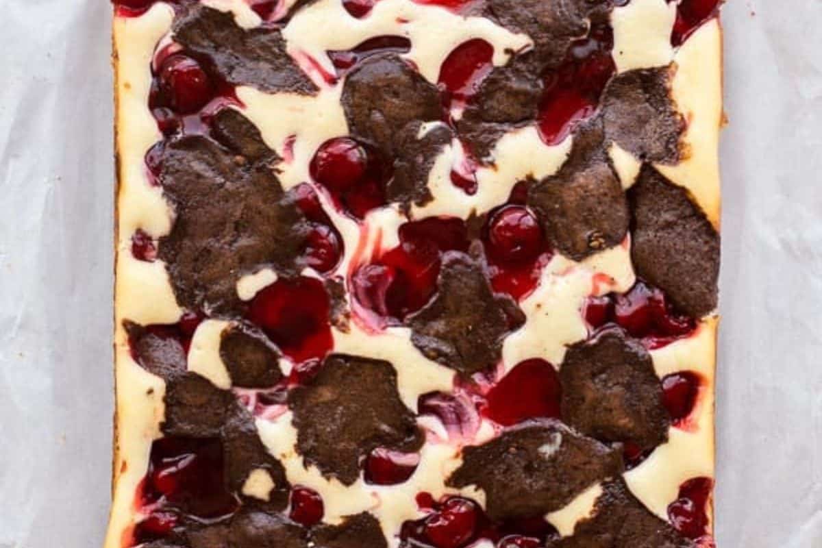 A rectangular cheesecake topped with chocolate brownie pieces and cherry filling, photographed from above on parchment paper—perfect inspiration for fresh cherry recipes.