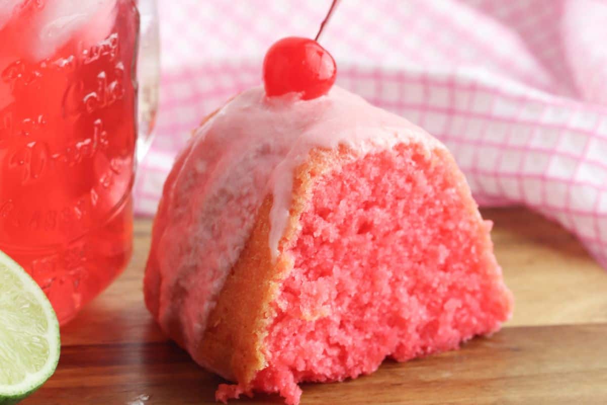A slice of pink bundt cake with white icing and a cherry on top, next to a glass of red drink and a lime slice on a wooden surface—a beautiful example of cherry desserts for anyone seeking fresh cherry recipes.