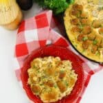 A red plate with a serving of Chicken Bacon Ranch Tater Tot Casserole sits on a red and white checkered cloth; a skillet with more casserole, fresh herbs, a pepper grinder, and a salt box are nearby.