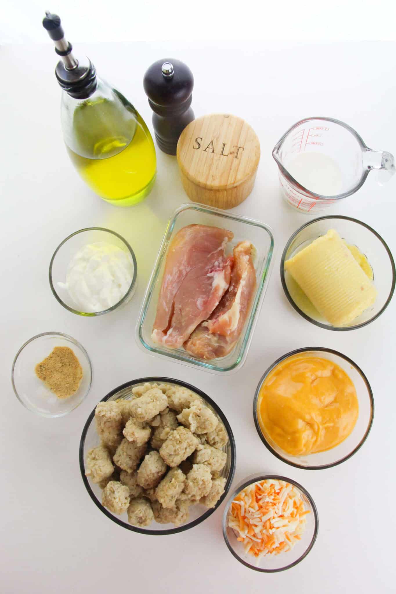 Overhead view of ingredients for Chicken Bacon Ranch Tater Tot Casserole, including olive oil, pepper grinder, salt, chicken breasts, butter, milk, Greek yogurt, bouillon, cheddar soup, shredded cheese, and sliced sausage.