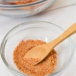 A small glass bowl containing Chicken Wing Seasoning Rub with a wooden spoon, placed in front of a larger bowl with seasoned food.