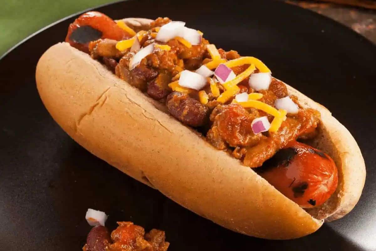 A grilled hot dog in a bun topped with chili, shredded cheese, and chopped onions on a dark plate—a true star spangled BBQ classic.