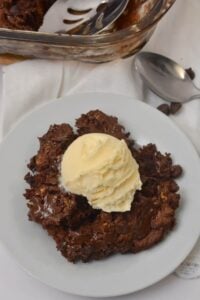 A serving of chocolate dump cake topped with a scoop of vanilla ice cream on a white plate, with a spoon and baking dish in the background.