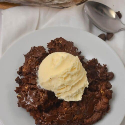 A serving of chocolate dump cake topped with a scoop of vanilla ice cream on a white plate, with a spoon and baking dish in the background.