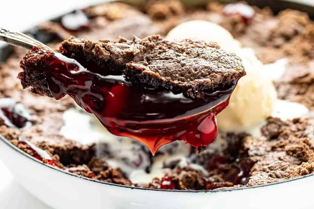 A close-up of a spoon lifting a serving of chocolate cherry cobbler—a tempting treat for fans of cherry desserts—with a scoop of vanilla ice cream melting in the background.