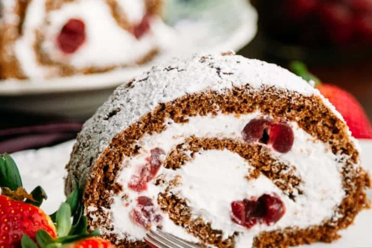 A close-up of a chocolate cake roll filled with whipped cream and fresh cherry, dusted with powdered sugar, with fresh strawberries on the side—a delightful treat for fans of fresh cherry recipes.
