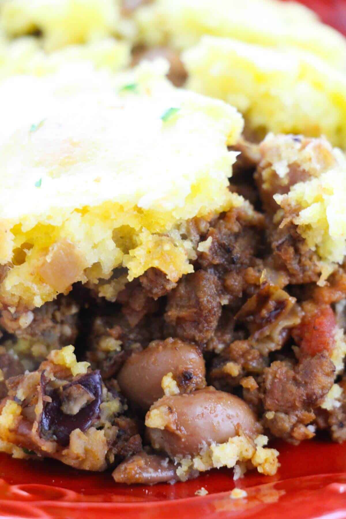 Close-up of a serving of tamale pie, similar to chili pie, with a cornbread topping, ground meat, beans, and vegetables on a red plate.