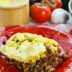 A serving of casserole topped with cornbread, garnished with parsley, on a red plate; fresh tomatoes, parsley, and kitchen items in the background.
