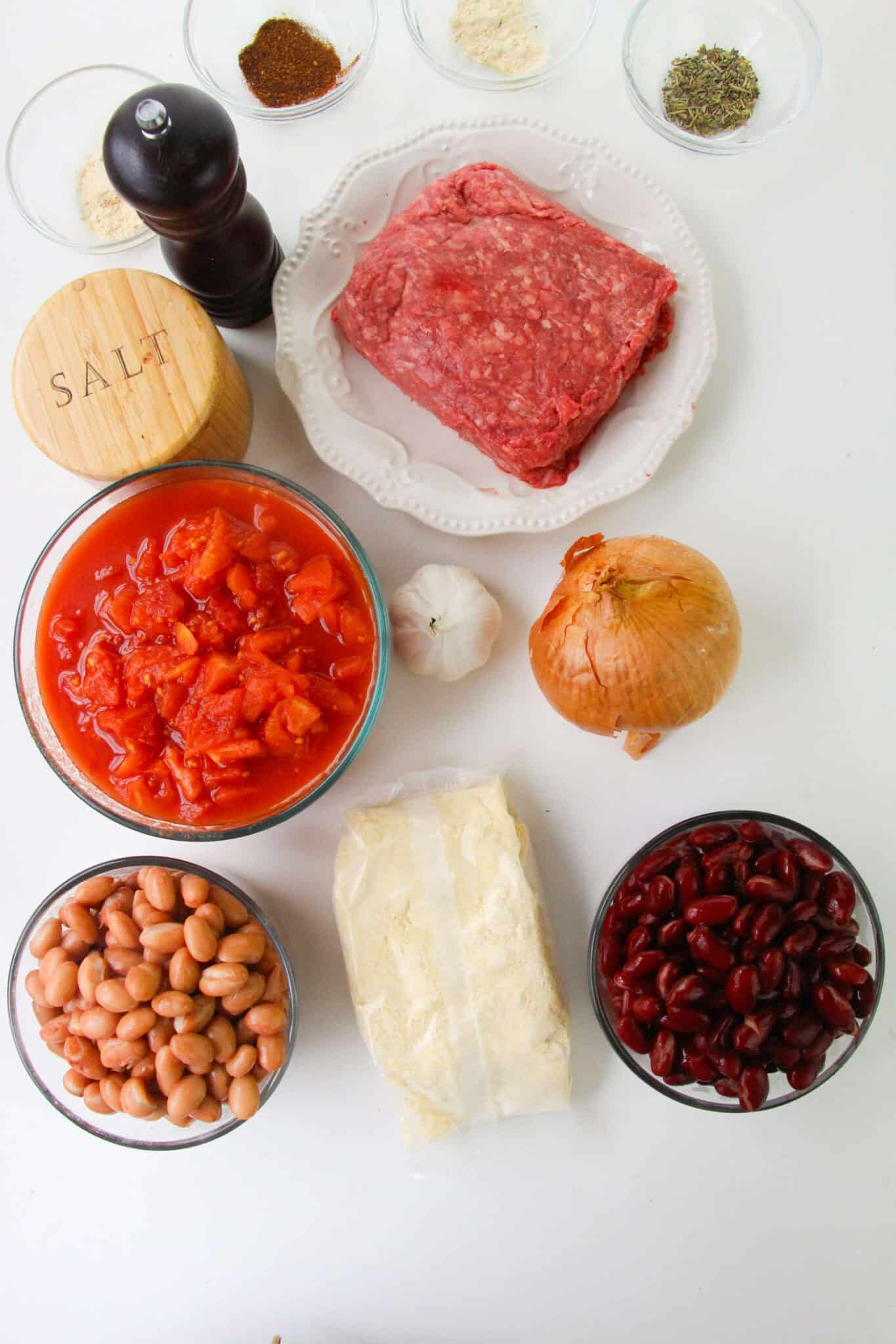 Ground beef, canned tomatoes, kidney beans, pinto beans, onion, garlic, spices, cornmeal, salt, and pepper are arranged on a white surface.