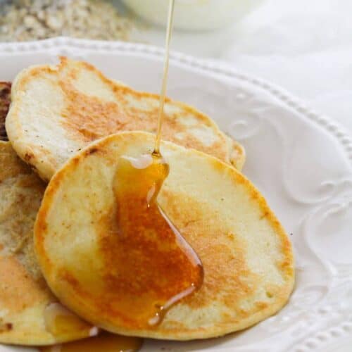 A close-up of breakfast on a white plate with syrup being poured on top; a bowl with a white mixture is visible in the background.
