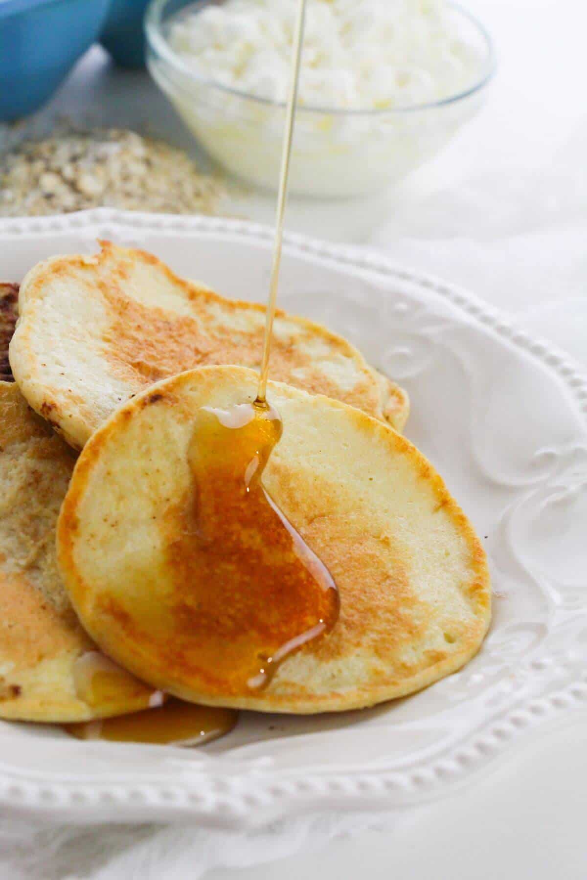 A close-up of breakfast on a white plate with syrup being poured on top; a bowl with a white mixture is visible in the background.