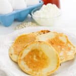Three flapjacks on a white plate, with a bowl of eggs, a glass container of syrup.