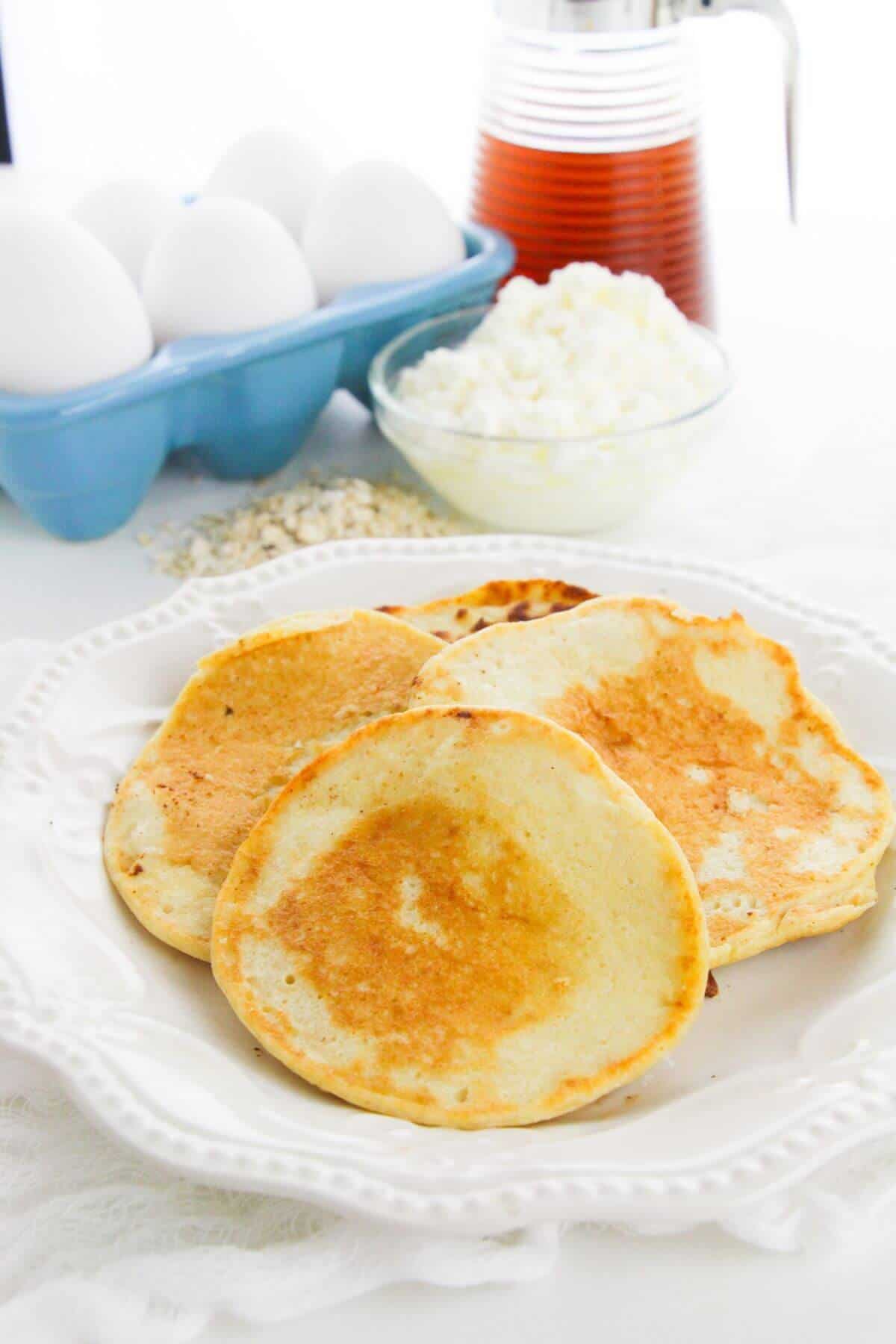 Three flapjacks on a white plate, with a bowl of eggs, a glass container of syrup.