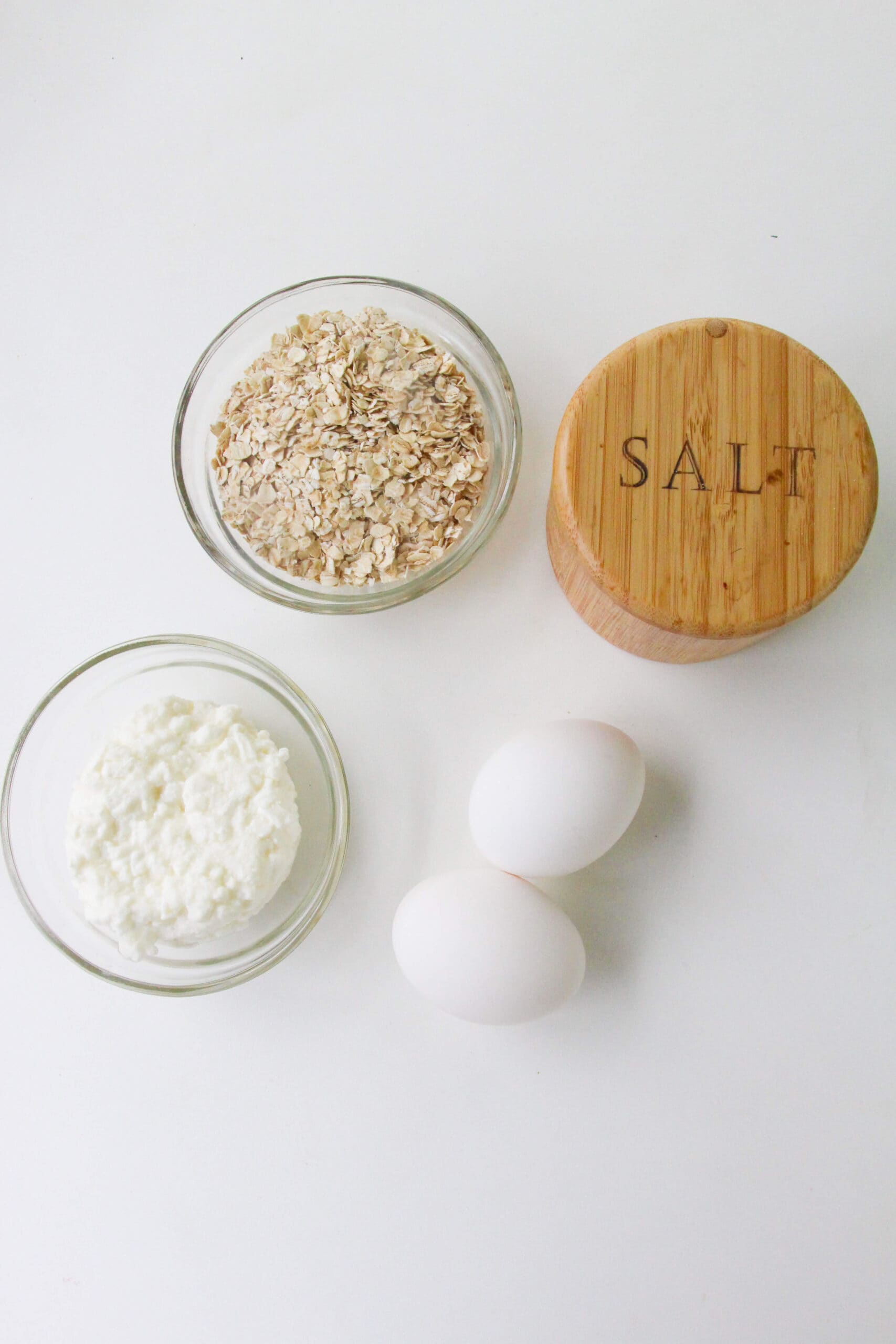 Two eggs, a small bowl of cottage cheese, rolled oats, and a wooden container labeled "SALT" on a white surface—perfect ingredients for delicious Cottage Cheese Oat Pancakes.