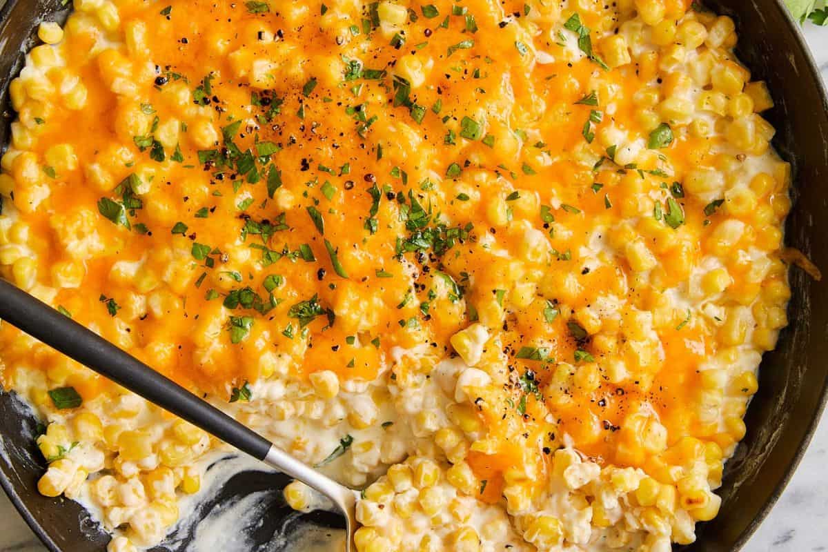 A skillet filled with creamy summer corn, topped with melted cheddar cheese, chopped herbs, and black pepper—perfect for enjoying delicious corn recipes on and off the cob. A serving spoon rests invitingly on the side.