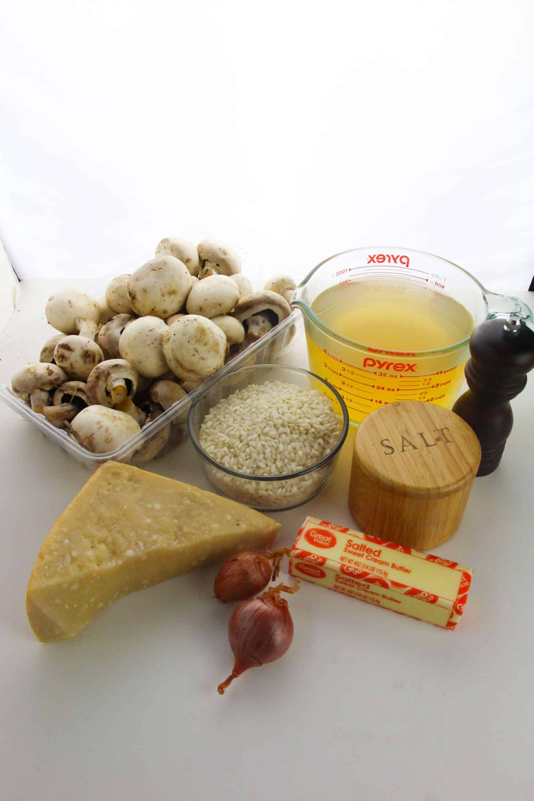 A display of Creamy Mushroom Risotto ingredients: white mushrooms, arborio rice, vegetable broth, Parmesan cheese, butter, shallots, salt, and a pepper grinder on a white surface.