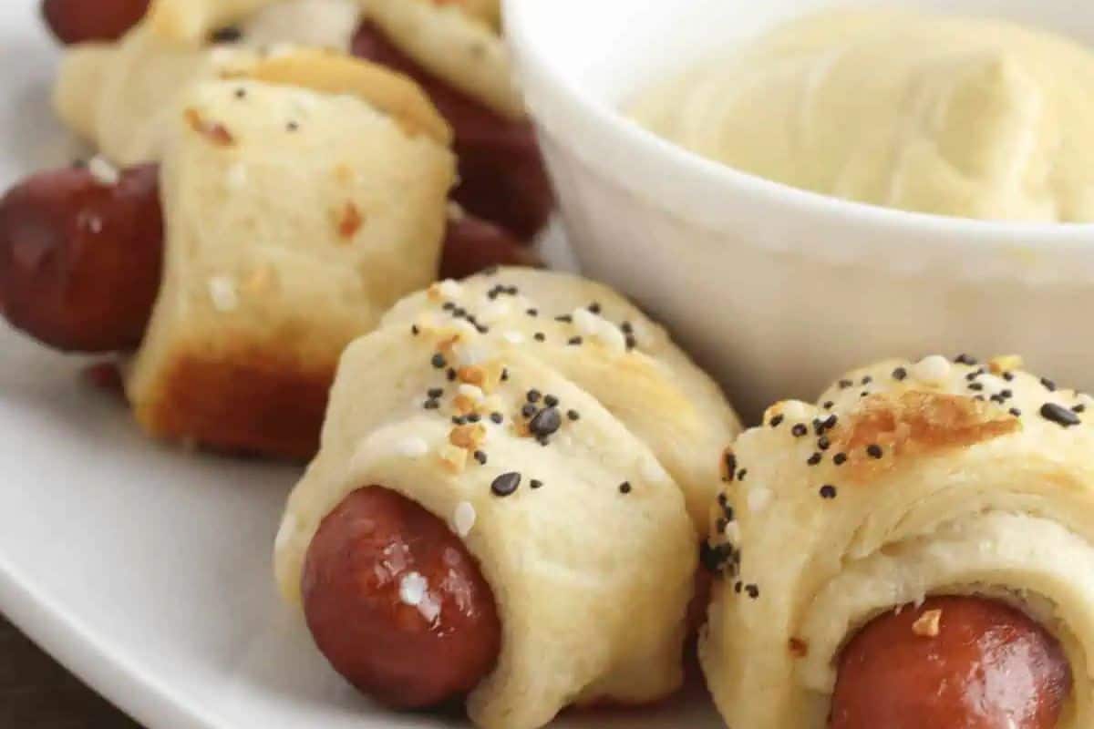 Mini sausages wrapped in baked crescent dough, sprinkled with seasoning, served on a white plate with a bowl of yellow dipping sauce—perfect for your Labor Day or Memorial Day gatherings and other patriotic party foods.