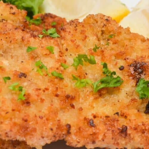 Close-up of breaded and fried pork cutlets, reminiscent of a classic pork cutlet, garnished with chopped parsley and served with lemon wedges on a white plate.