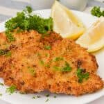 Two thinly pounded and breaded meat garnished with chopped parsley and served with lemon wedges and parsley sprigs on a white plate.