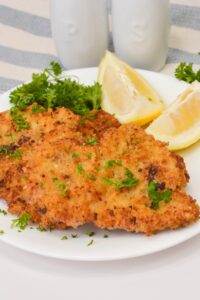 Two thinly pounded and breaded meat garnished with chopped parsley and served with lemon wedges and parsley sprigs on a white plate.