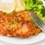 A plate with breaded cutlet, steamed broccoli, lemon wedges, and a fork resting on the side.