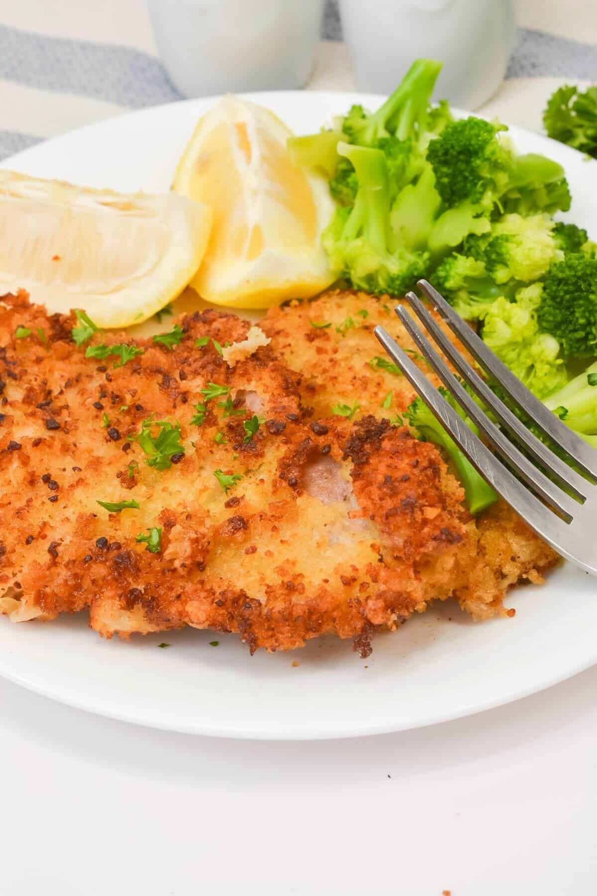 A plate with breaded cutlet, steamed broccoli, lemon wedges, and a fork resting on the side.