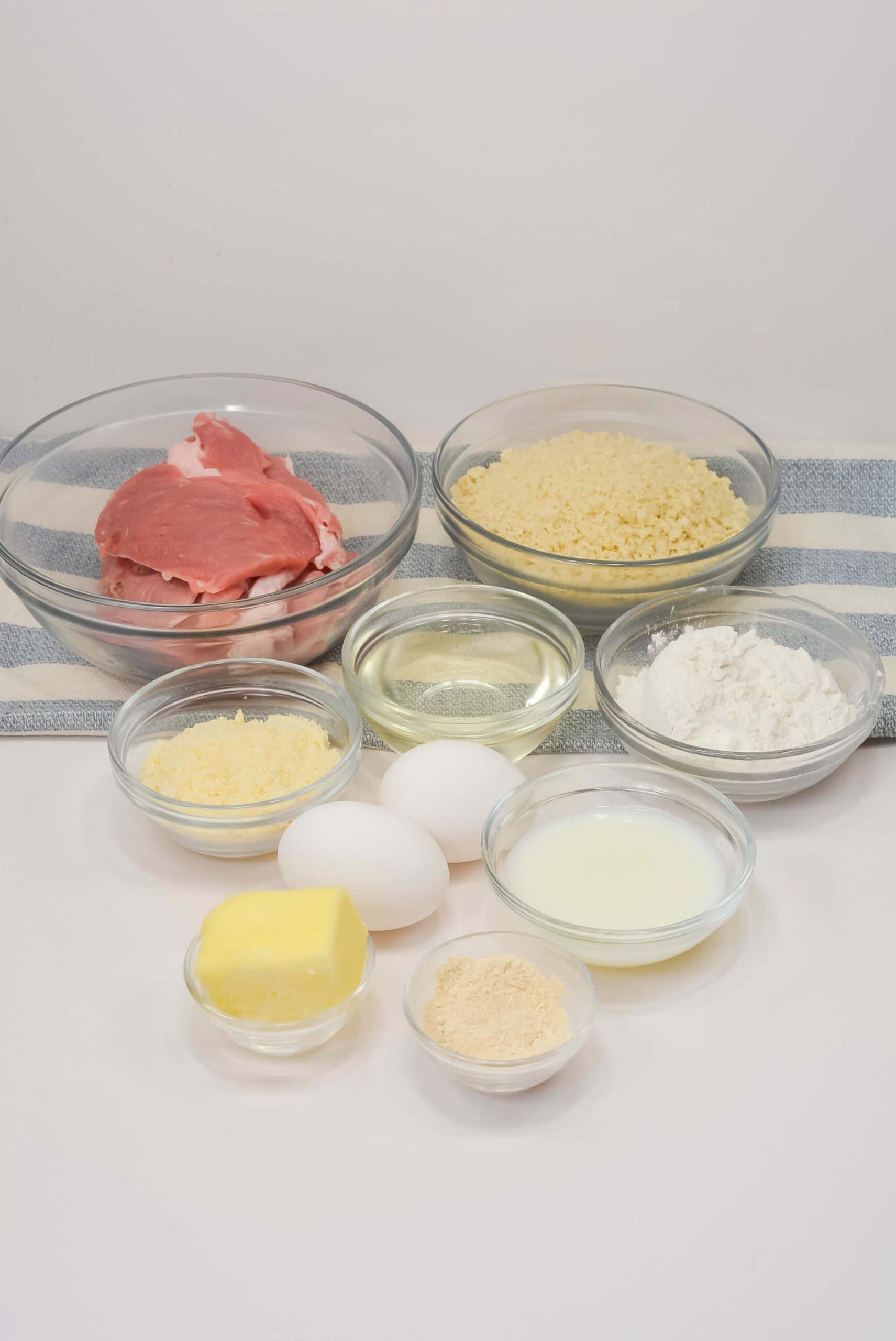 Assorted ingredients in glass bowls, including raw pork cutlet, eggs, flour, grated cheese, breadcrumbs, oil, butter, milk, and a light-colored powder, arranged on a striped towel.
