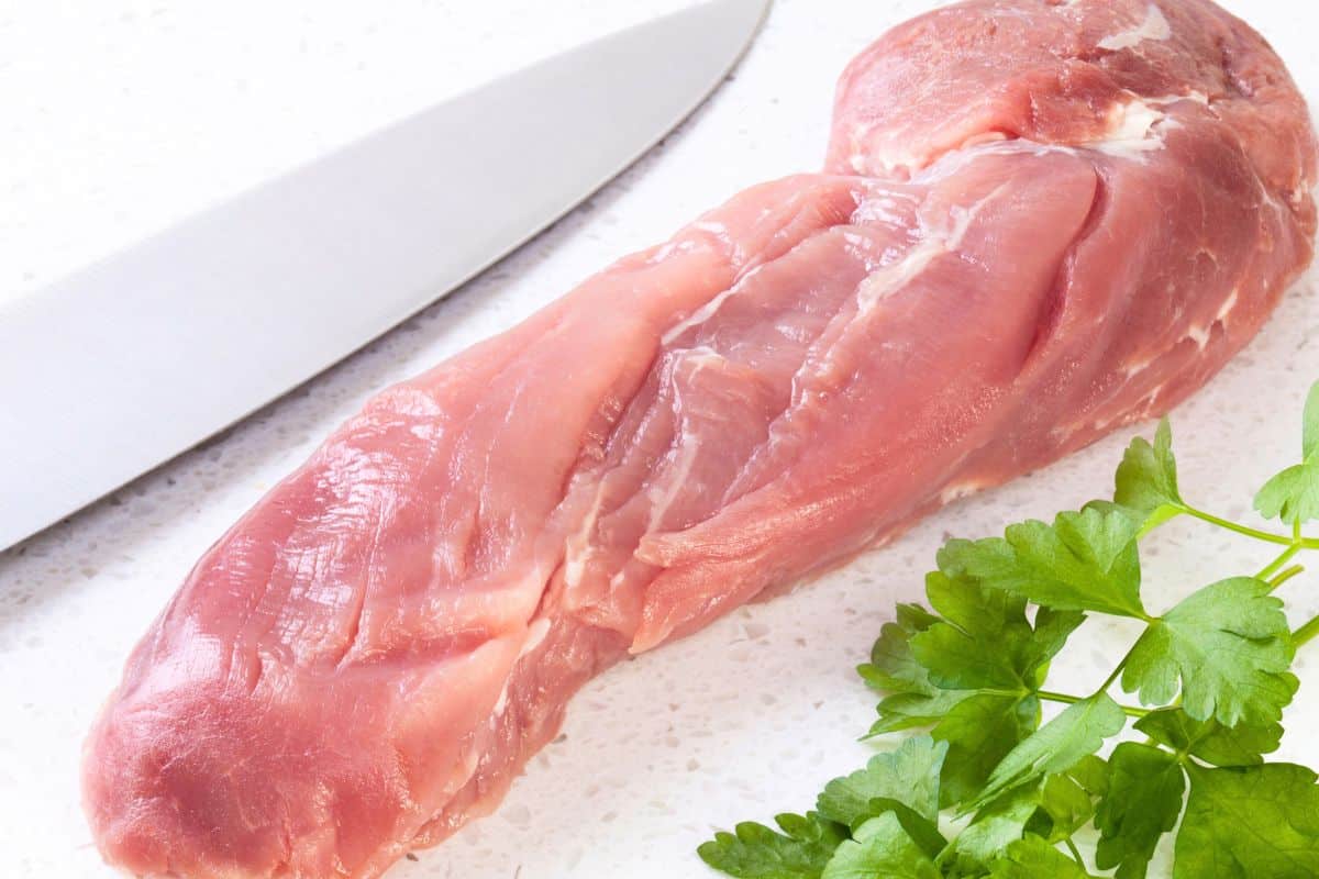 Raw pork tenderloin on a white surface next to a kitchen knife and a sprig of fresh parsley, ideal for slicing into tender pork cutlets.