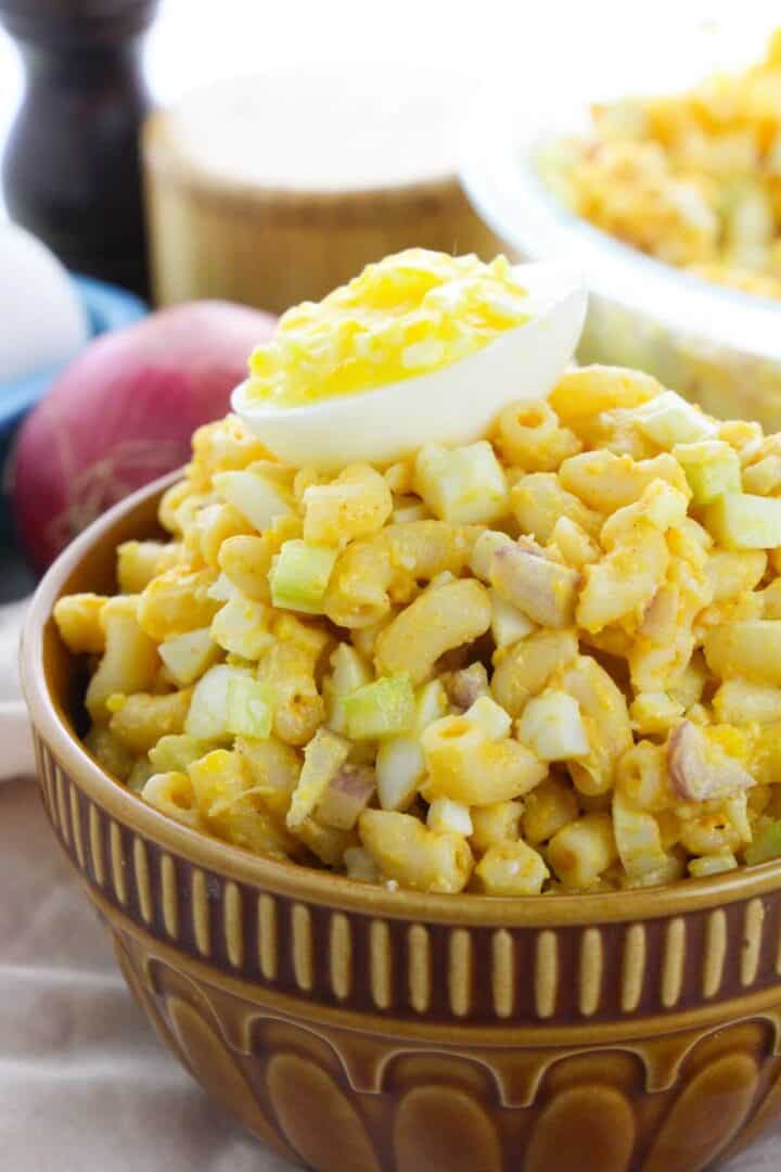 A bowl of Deviled Egg Macaroni Salad with chopped celery and egg, topped with half a boiled egg, sits on a table with an onion and whole eggs in the background.