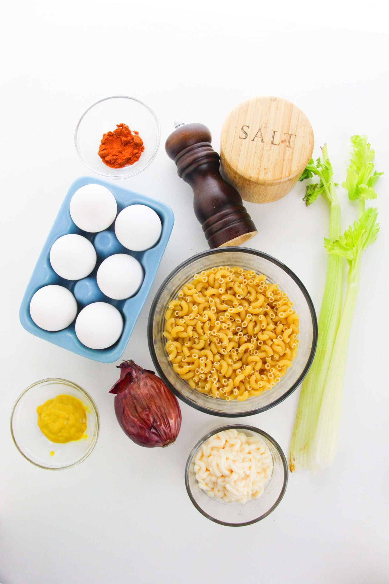 Ingredients for the dish including eggs, elbow macaroni, celery stalks, red onion, mustard, paprika, salt, pepper, and mayonnaise—arranged neatly on a white surface.