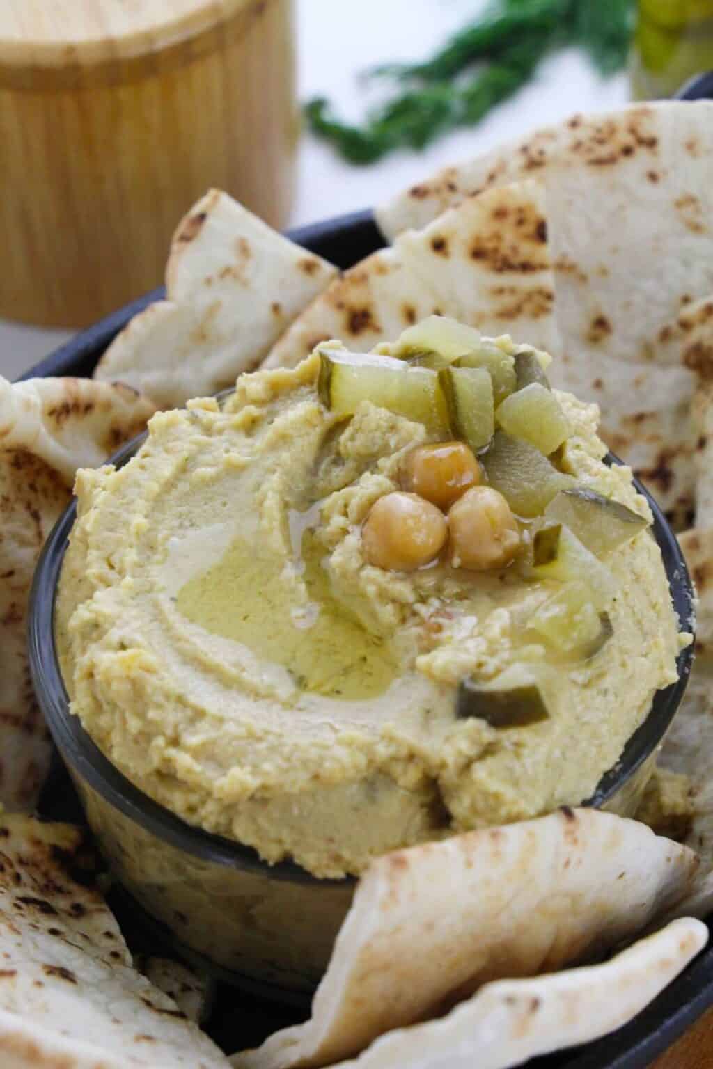 A bowl of dill pickle hummus topped with chopped pickles, whole chickpeas, and olive oil, surrounded by pieces of pita bread.