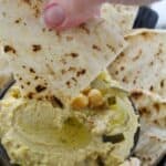 A hand dips a piece of grilled flatbread into a bowl of dip garnished with whole chickpeas and olive oil. Other pieces of flatbread are arranged around the bowl.