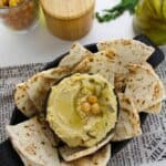 A bowl of Dill Pickle Hummus topped with chickpeas and pickles, surrounded by pita bread slices, sits on a tray with a napkin, salt container, and chickpeas in the background.