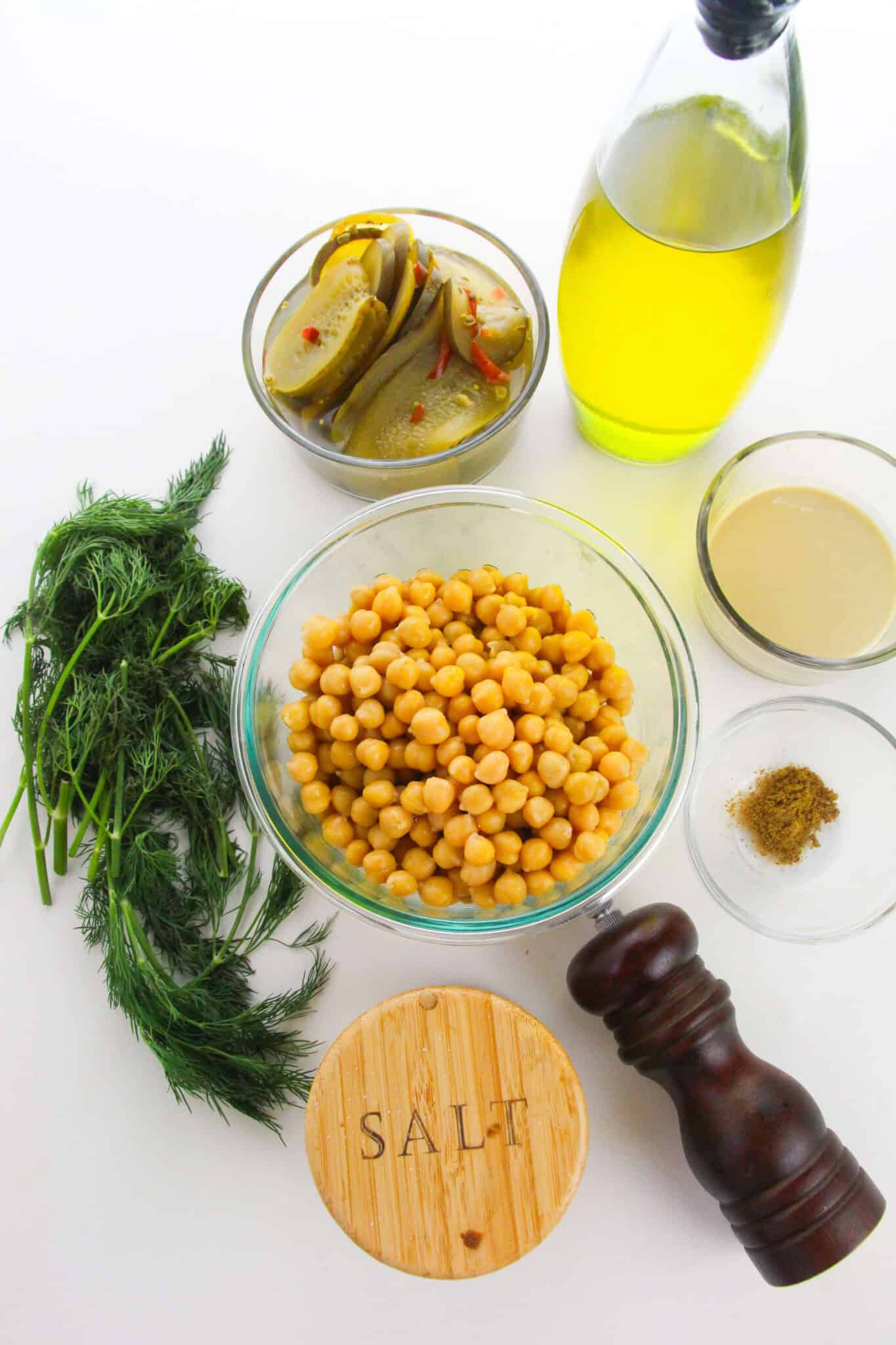 Bowls of pickles, olive oil, tahini, chickpeas, dill, ground spices, and salt—everything you need to whip up fresh Dip—are artfully arranged on a white surface with a pepper grinder nearby.