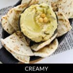 A bowl of creamy Dill Pickle Hummus topped with chickpeas and pickles, surrounded by pieces of pita bread on a black plate.