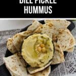 A bowl of creamy Dill Pickle Hummus topped with chickpeas and chopped pickles, surrounded by grilled pita bread slices on a black platter.